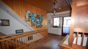 Una escalera de madera con barandilla, un tablón de anuncios con folletos de viajes, un radiador y una ventana con cortinas estampadas.
