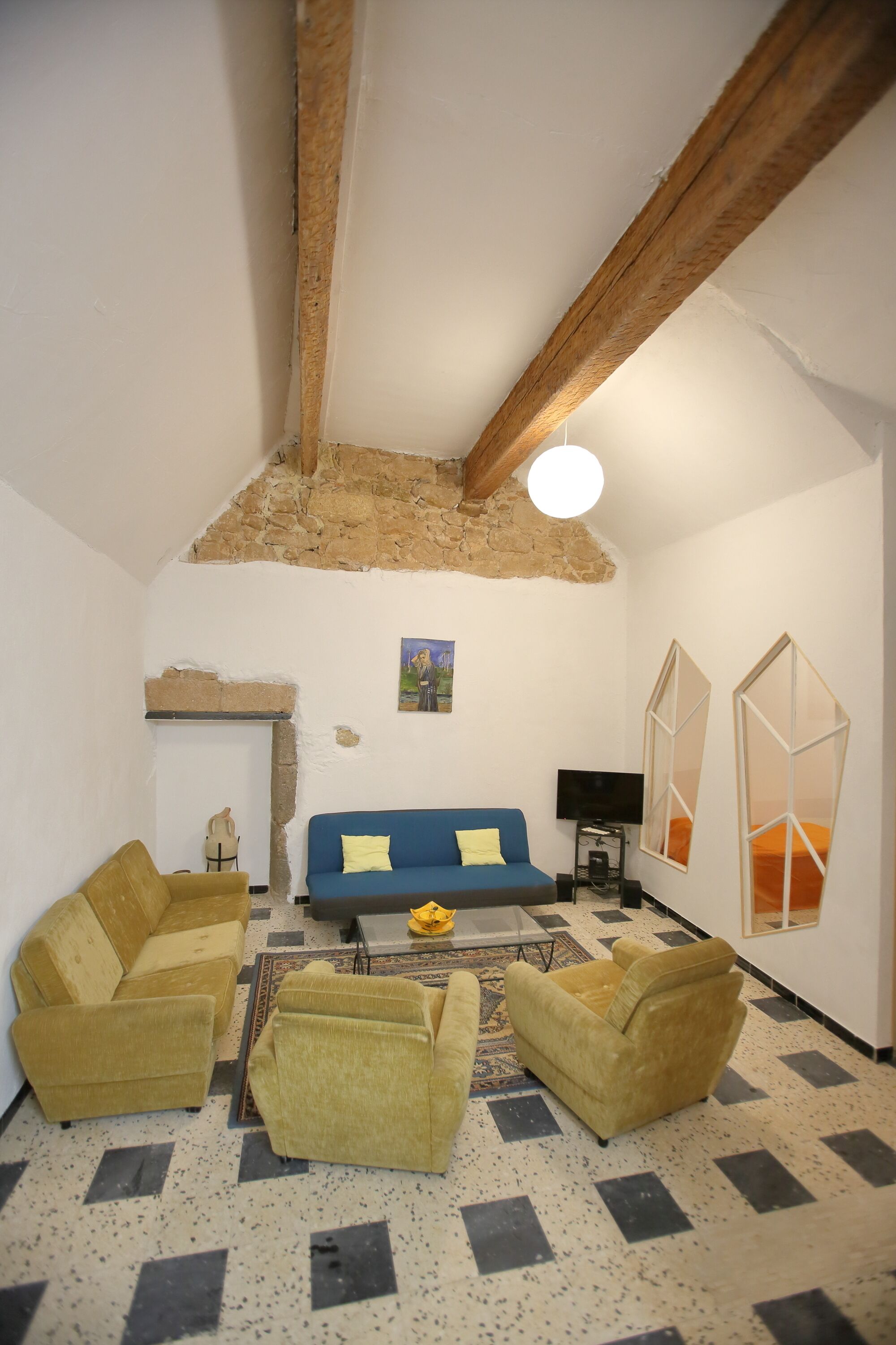Un salon avec un canapé bleu, des coussins jaunes et un fauteuil jaune. On y trouve une télévision et une petite table sur laquelle est posé un vase. La pièce est dotée d’un haut plafond avec des poutres apparentes et d’un mur en pierre.