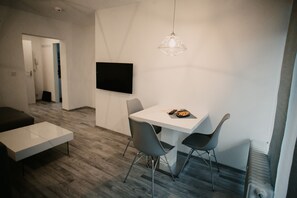 A modern, minimalist room with a white table, two grey chairs, a white sofa, and a mounted television.