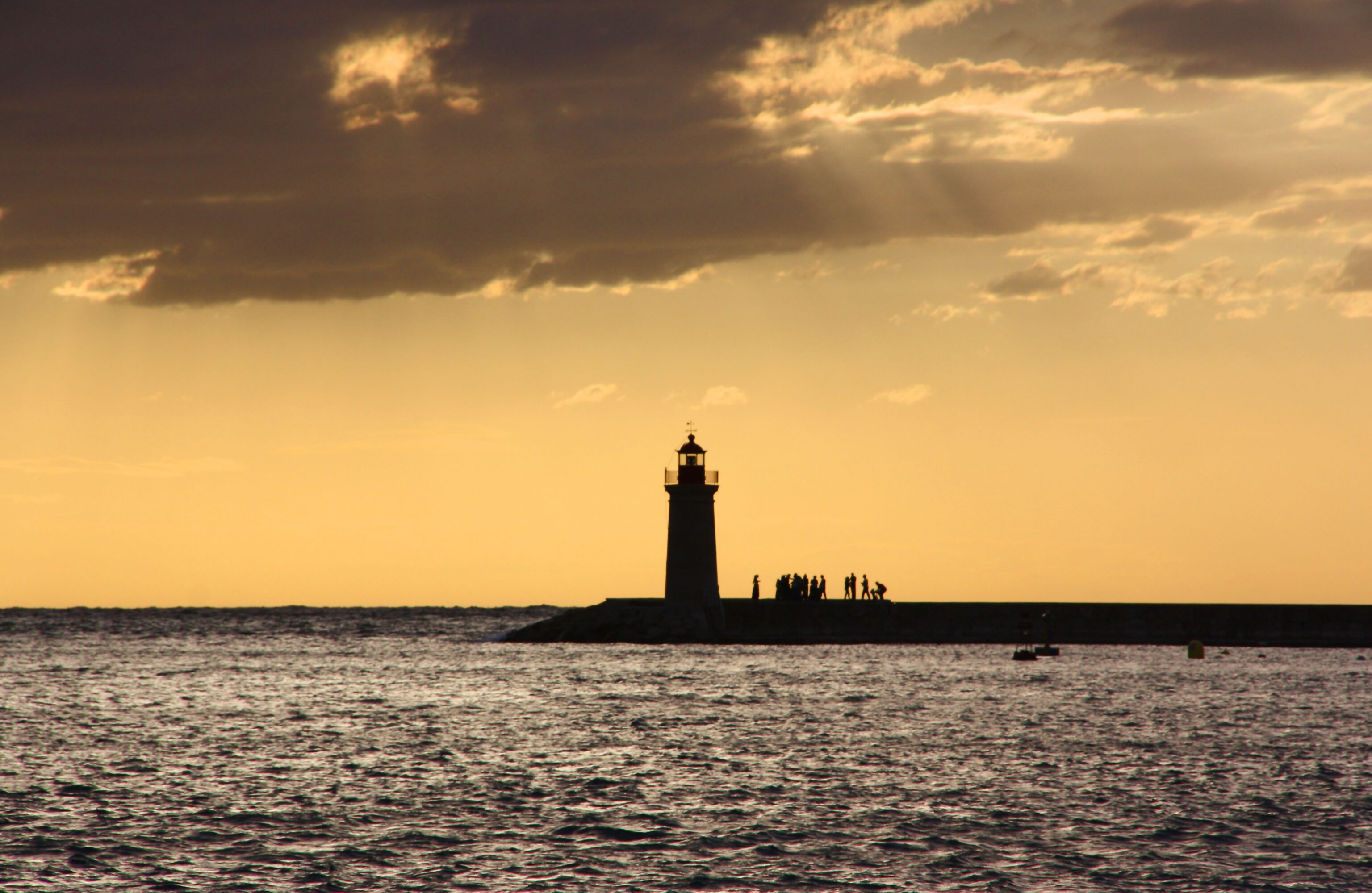 Silueta de un faro contra un cielo al atardecer, con mar tranquilo y un muelle con gente.