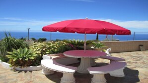 Eine Terrasse mit einem runden Tisch und Bänken, umgeben von Topfpflanzen und einem roten Sonnenschirm.