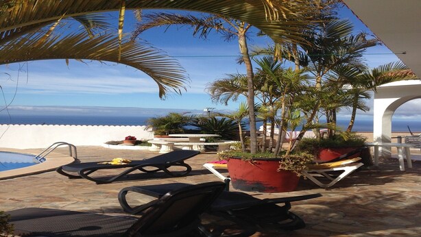 Ein Poolbereich mit Liegestühlen, einem roten Pflanzgefäß mit Pflanzen und Blick aufs Meer.