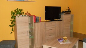 Una sala de estar con televisor, armarios de madera, una mesita con revistas, una planta y un reloj en la pared.