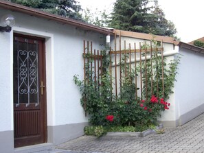 Uma casa com fachada branca, porta marrom e um pequeno jardim com flores vermelhas.