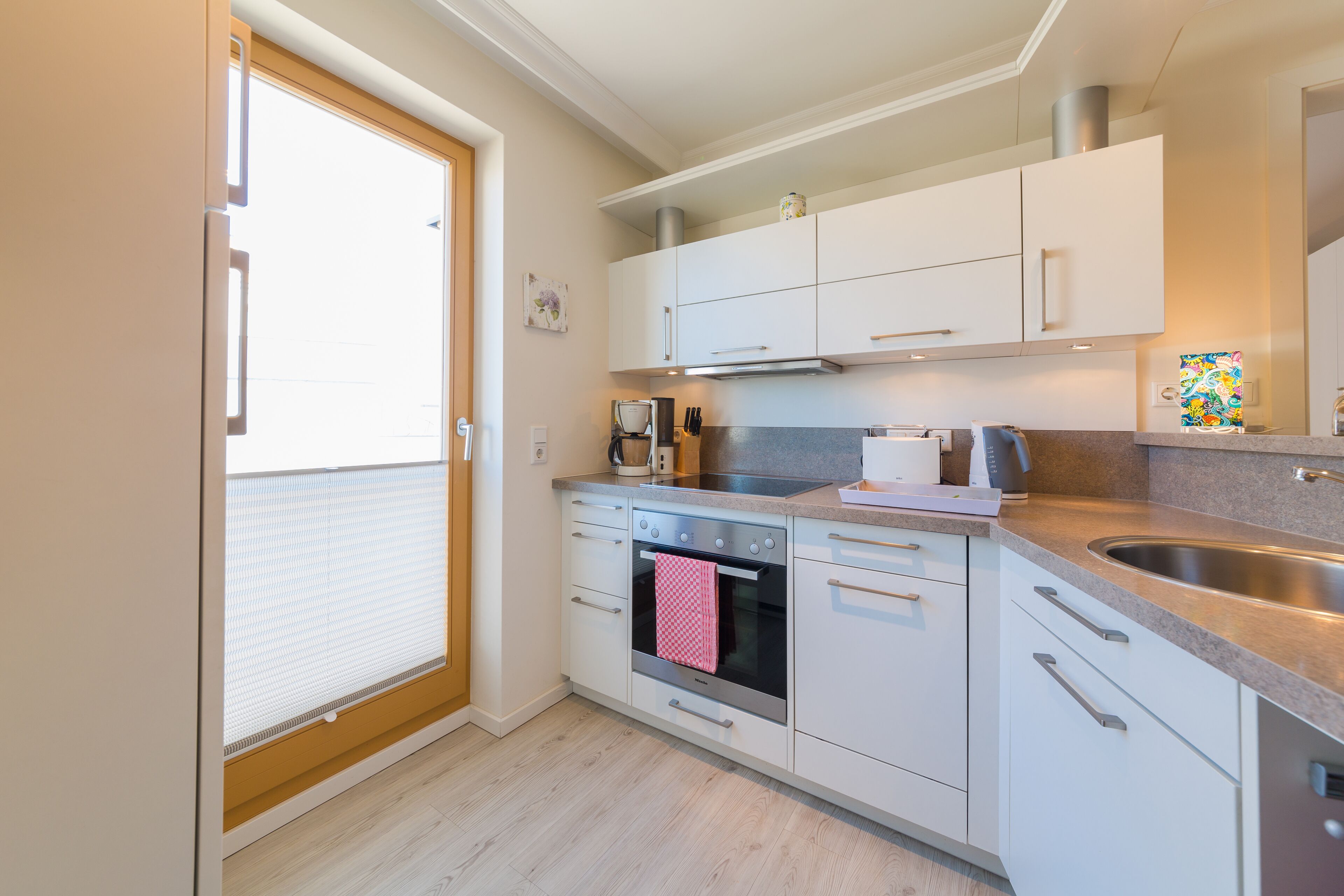 Een moderne keuken met witte kastjes, een inbouwoven, een roestvrijstalen spoelbak en een raam met zonwering.