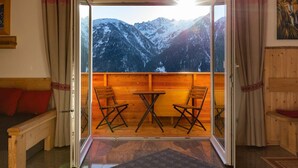 Una habitación con un banco de madera, una mesa y dos sillas orientadas hacia una vista de montaña a través de una puerta abierta.