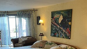 A spacious bedroom with a large bed, a wooden headboard, a painting on the wall, and a view through a glass door.