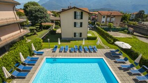 A rectangular swimming pool with blue water, surrounded by lounge chairs and umbrellas, set against a backdrop of residential buildings and mountains.