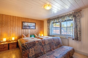 A wooden-paneled room with a bed, two bedside tables with lamps, a window with curtains, and a painting on the wall.
