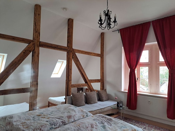 A room with exposed wooden beams, a bed with floral bedding, a small table, and a window with red curtains.