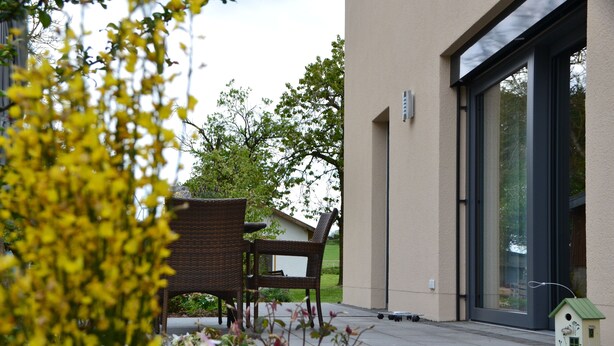A patio area with a wicker chair, a small birdhouse, and a building with large windows. - Eifelgold - Comfortable, spacious 5-star apartment in a quiet location  (Habscheid)