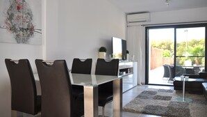 A modern dining area with a glass table and black chairs, a flat-screen TV, and a view of the outdoors through large sliding doors. - Modern bungalow with heated pool, 50m from the sea - whirlpool - (Cala Pi)