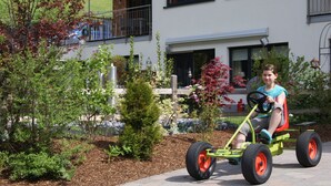 Un niño montando un triciclo pequeño verde con ruedas rojas en un jardín con varias plantas y un edificio al fondo.