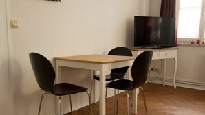 A room with a wooden floor, a white table with two black chairs, a television on a small white table, and a window with curtains.