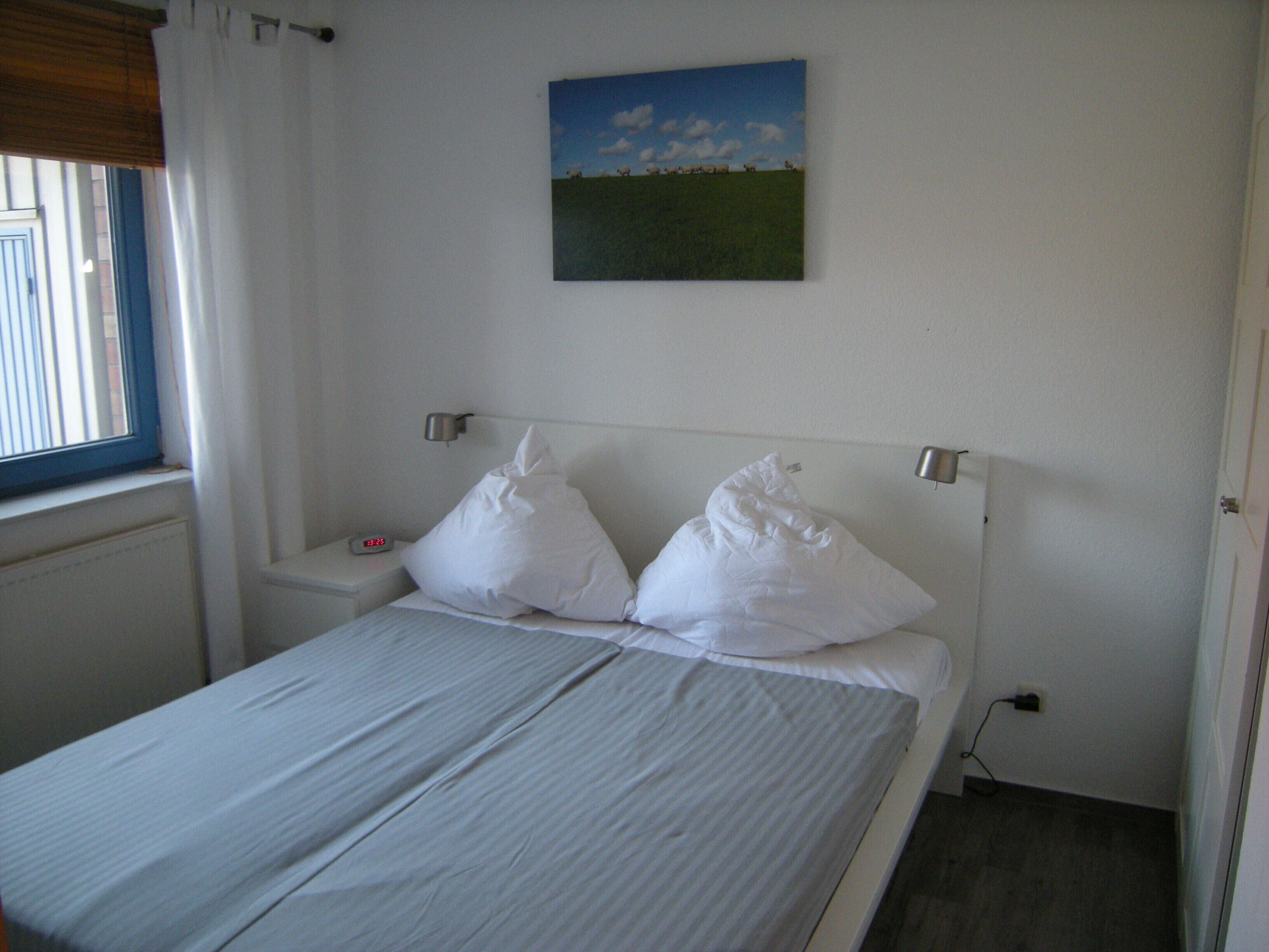 A bedroom with a bed, two pillows, a bedside table with a clock, a wall-mounted lamp, and a painting of a field.
