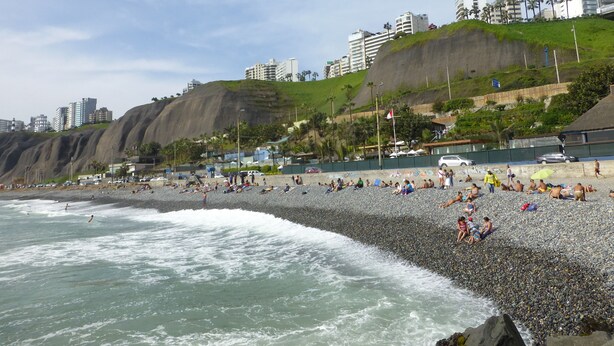 A crowded pebble beach with waves crashing onto the shore, a grassy hill with buildings in the background, and a clear sky. - MALECÓN BALTA: best location, friendly service, clean & comfortable, safe (Miraflores)