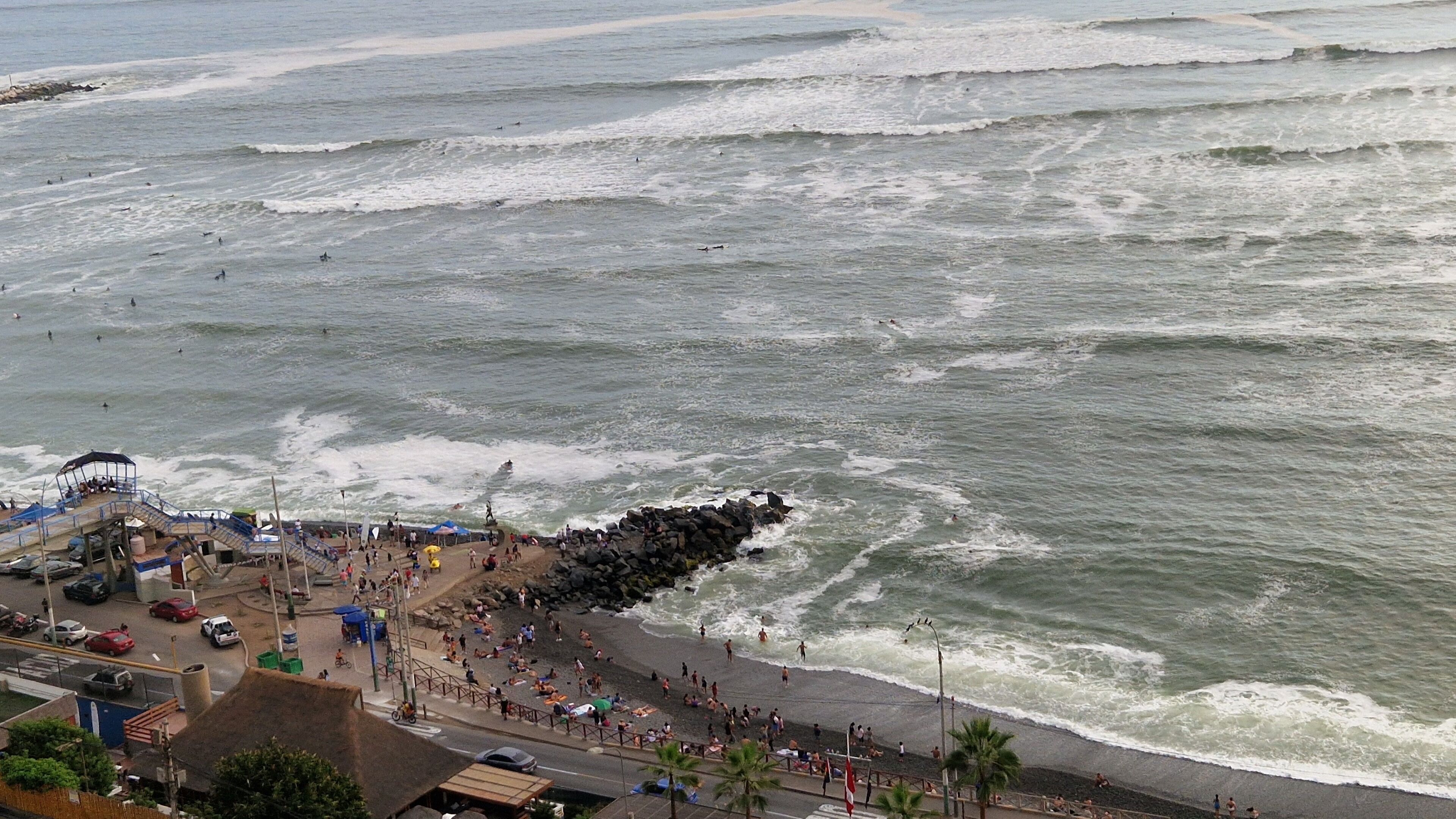 A beach with a rocky breakwater, people sunbathing, and a pier with umbrellas.
