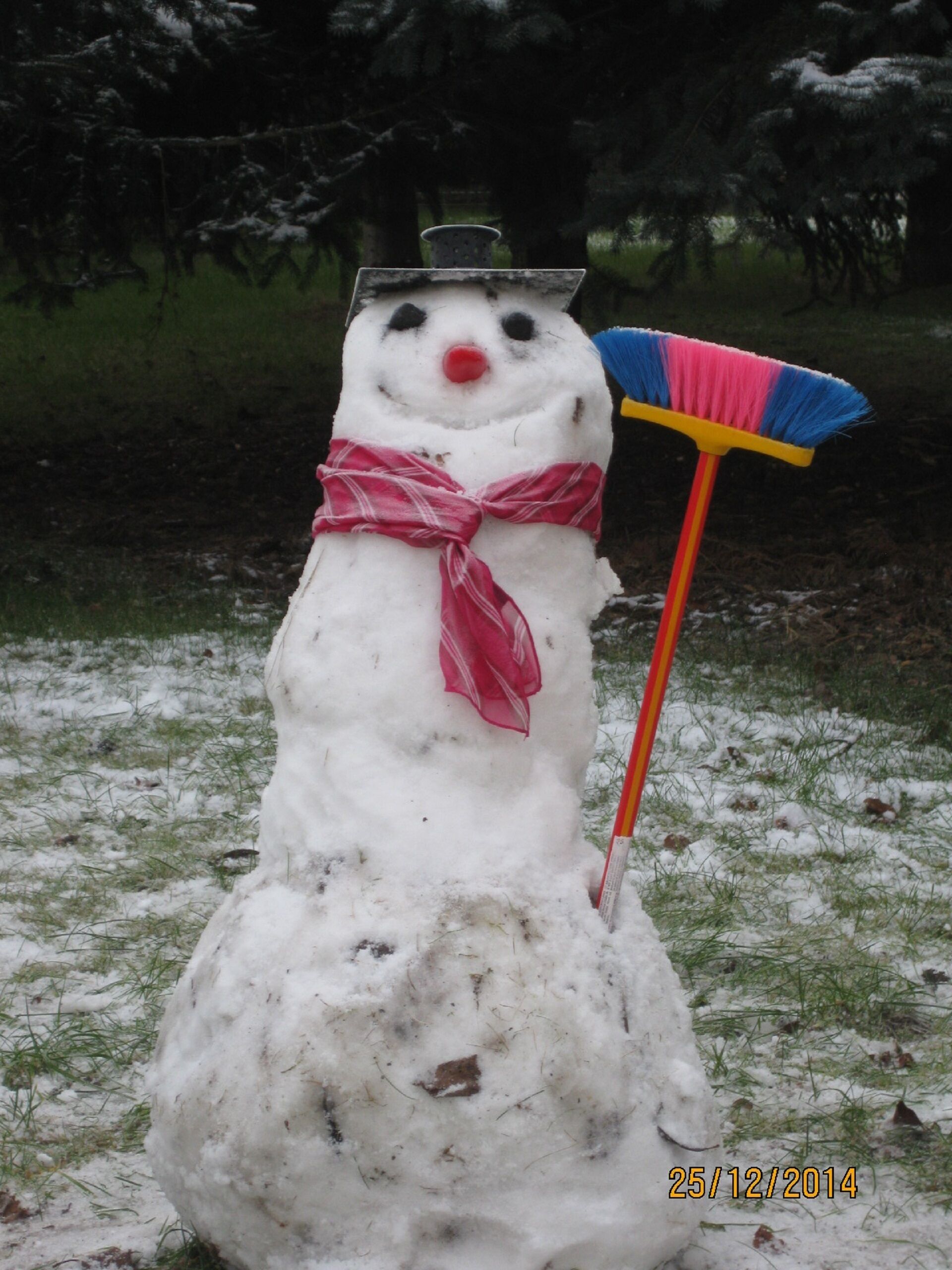Ein Schneemann mit Besen und rotem Schal, stehend auf einer schneebedeckten, grasbewachsenen Fläche.