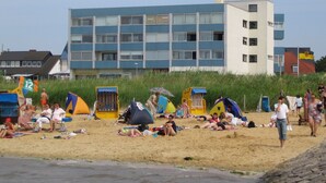 Une plage où des gens se prélassent au soleil et où l’on voit des tentes. Un immeuble à plusieurs étages, doté de balcons, se trouve à l’arrière-plan.