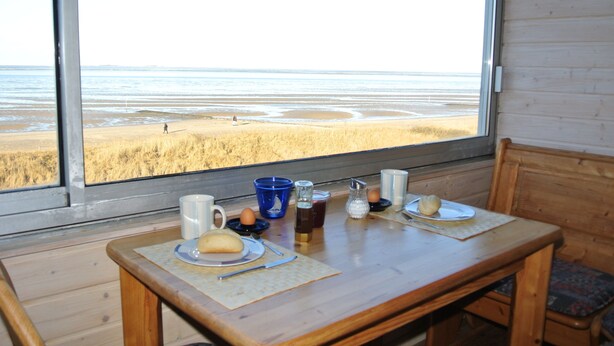Une table en bois, dressée pour le petit-déjeuner, avec vue sur une plage par la fenêtre.