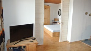 Une pièce avec un plancher en bois, un lit avec un matelas, un téléviseur posé sur un meuble en bois et une porte donnant sur une autre pièce.