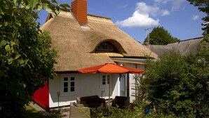 Ein traditionelles Haus mit einem strohgedeckten Dach, einem roten Schornstein und einer kleinen Veranda mit Korbmöbeln.
