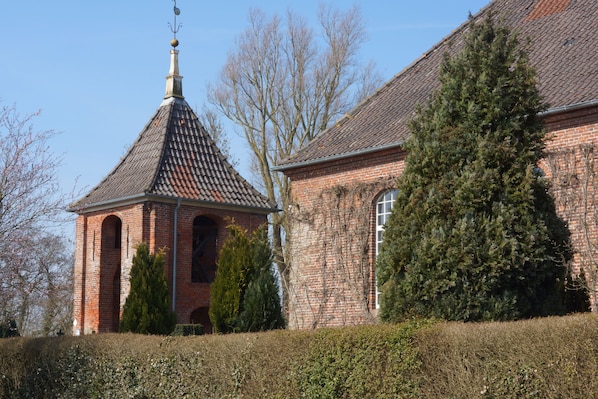 A brick building with a conical roof and a bell tower, surrounded by trees and a hedge.