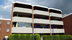 A multi-story residential building with balconies, a brick facade, and a parking area.