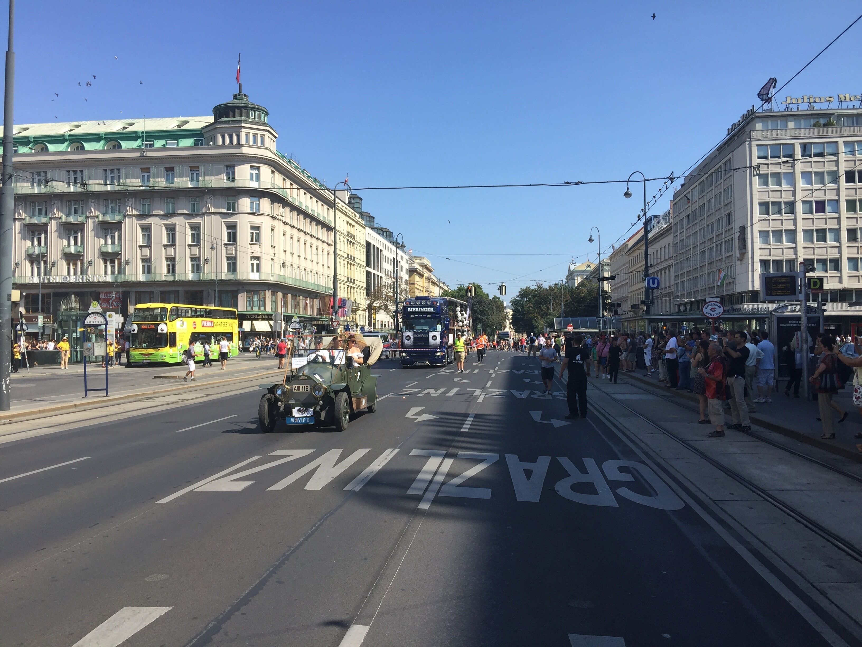 Eine Straße mit einer Straßenbahn, einem Oldtimer und einer Menschenmenge.