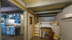 Une petite pièce comprenant un lit superposé, un petit bureau avec une chaise, une kitchenette équipée d’une cuisinière et d’un évier, et un plafond en bois.