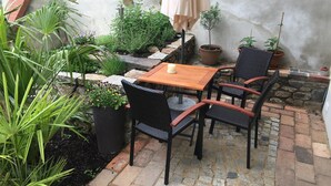 Un espace patio avec une table en bois et quatre chaises en osier, entouré de plantes en pot et d’un mur en pierre.