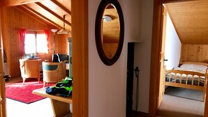 A cozy room with a wooden ceiling, a bed, a desk with a laptop, wicker chairs, and a red rug.