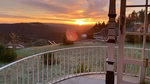 A balcony with a view of a sunset over a valley, a wooden railing, and a window with a thermometer. - Large cozy apartment centrally in Winterberg with panoramic views (Winterberg)
