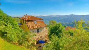 Una casa in pietra con tetto in tegole, immersa nel verde e con montagne sullo sfondo.
