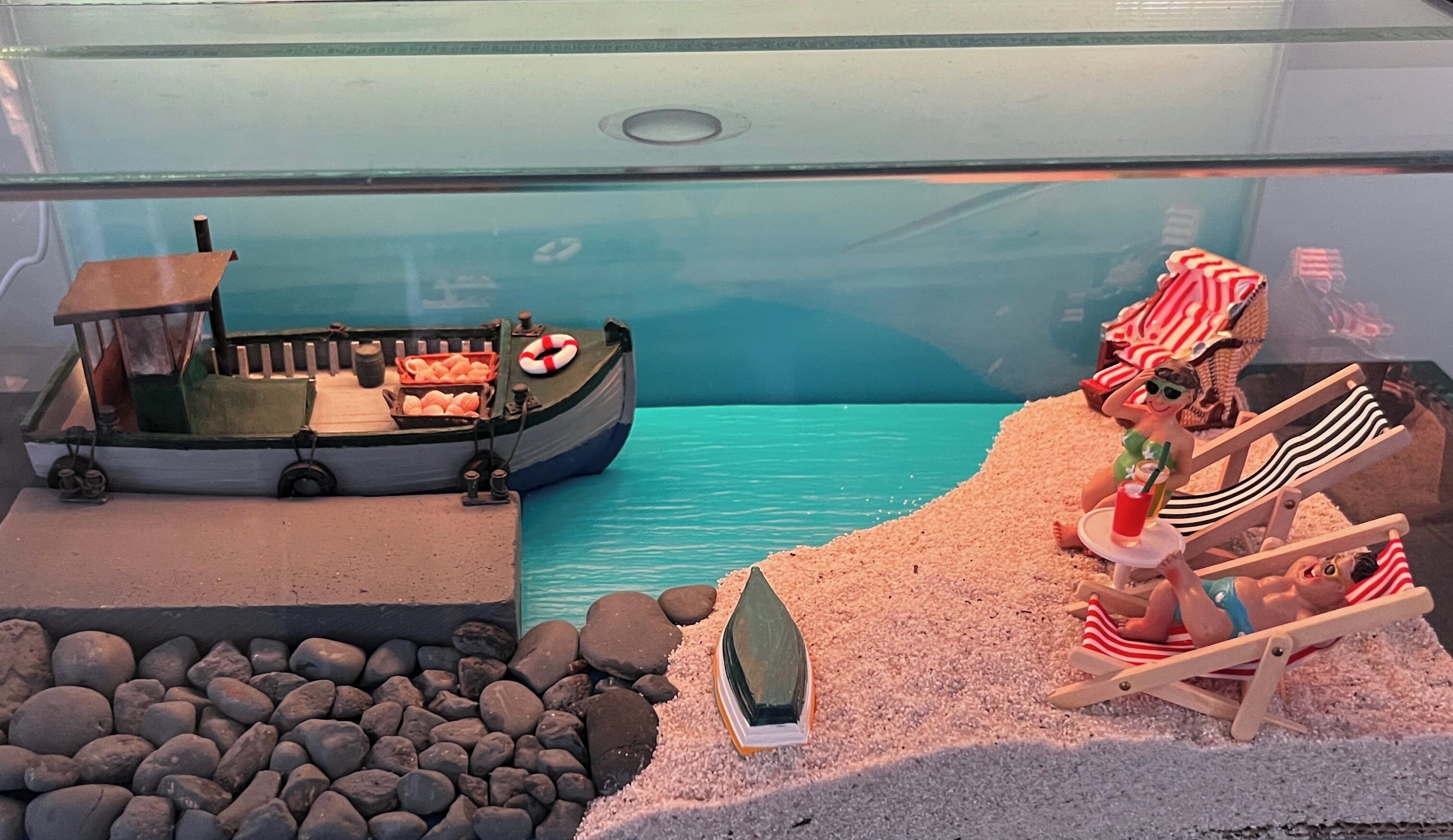 A model of a boat with a cabin and a lifebuoy, a sandy beach with rocks, and a figure in a striped beach chair.