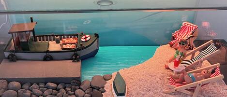 A model of a boat with a cabin and a lifebuoy, a sandy beach with rocks, and a figure in a striped beach chair.