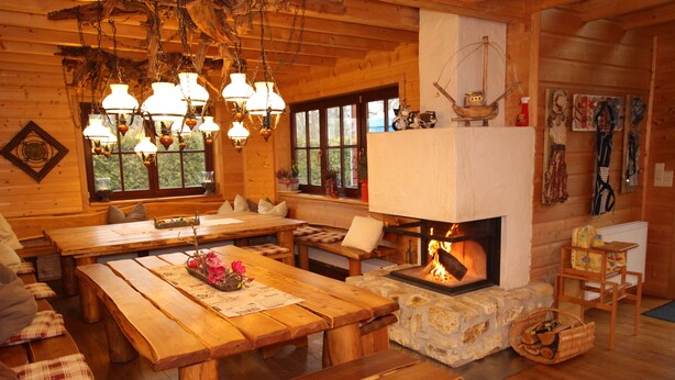 A cozy cabin interior with a large wooden dining table, benches, a fireplace, and a stone fireplace surround.