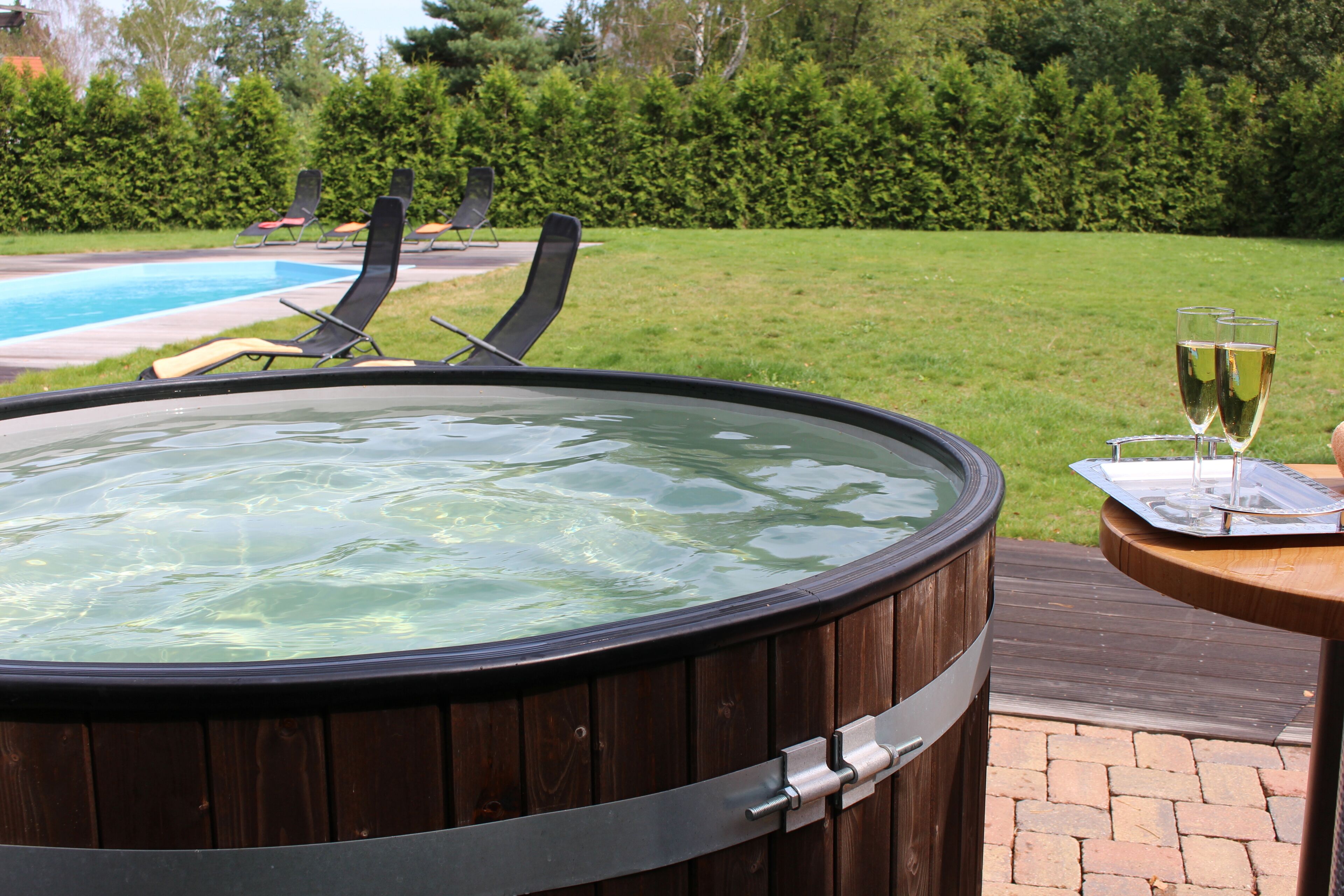A hot tub with a wooden exterior, a pool in the background, and a grassy area with lounge chairs.