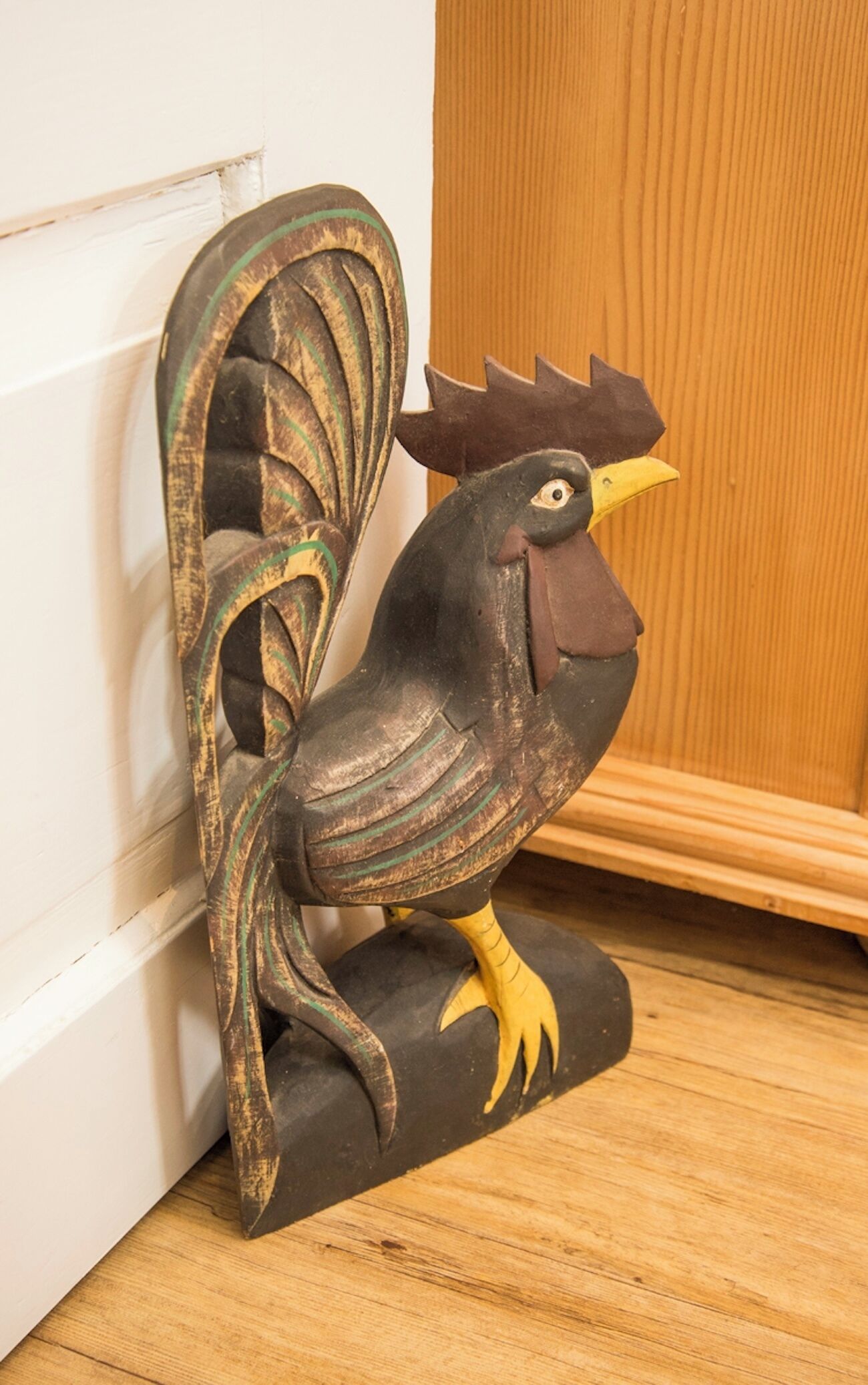 Una escultura rústica de gallo de madera sobre un piso de madera, junto a una puerta blanca.