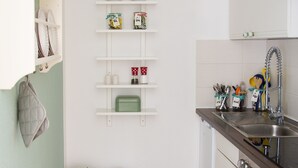 Una cocina compacta con armarios blancos, un lavamanos y piso de baldosas. Hay dos botes de basura y un recipiente verde en el suelo.