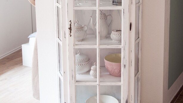 Un mueble blanco de estilo antiguo con varios estantes, algunos con cristalería y objetos de cerámica.