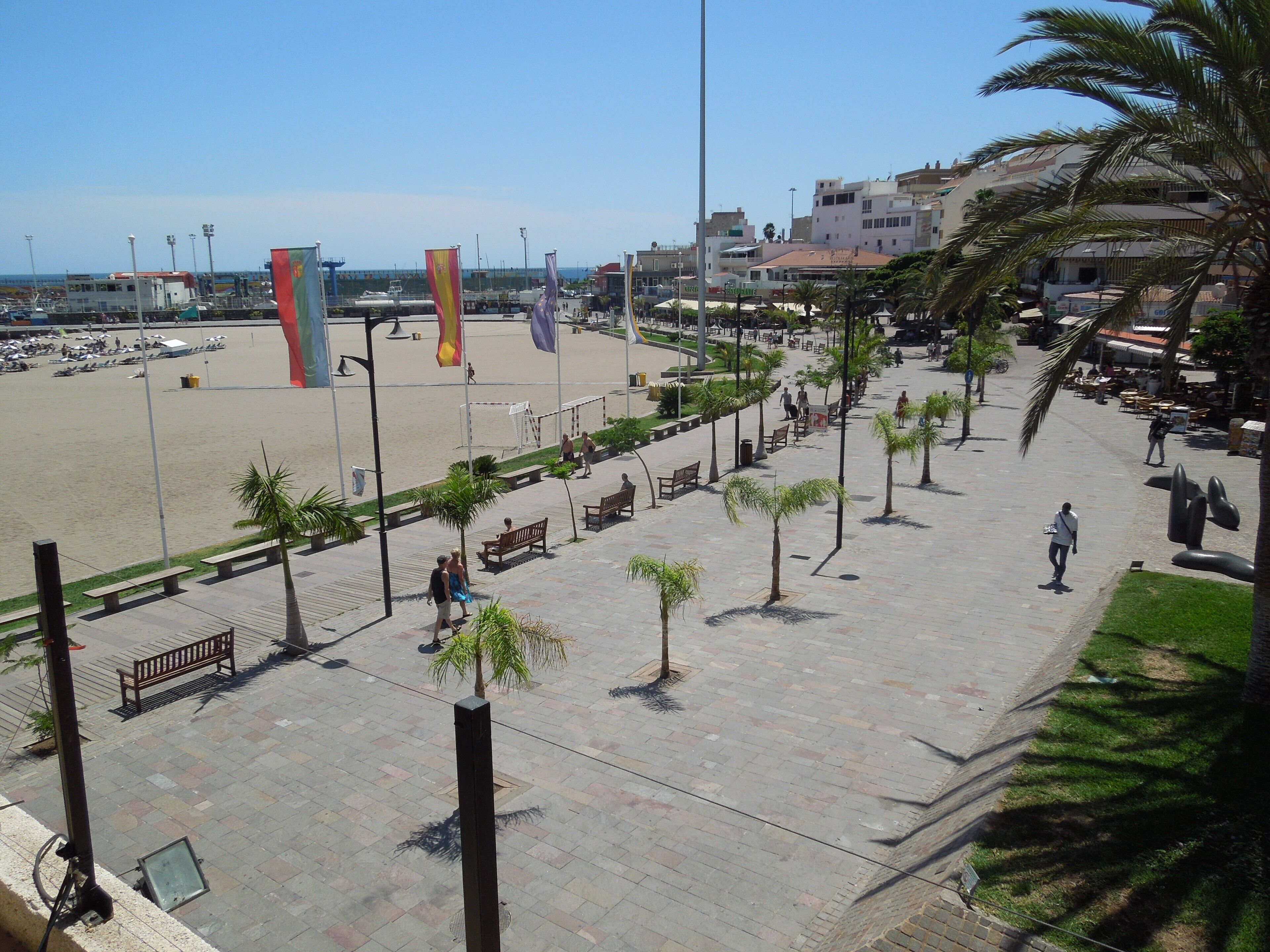 A coastal promenade with a sandy beach, benches, palm trees, and a clear sky.
