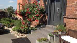A residential entrance with a wooden deck, a red flowering bush, and a bench. - Quiet situated bungalow with a terrace overlooking the water and in nature (Garz/Rügen)