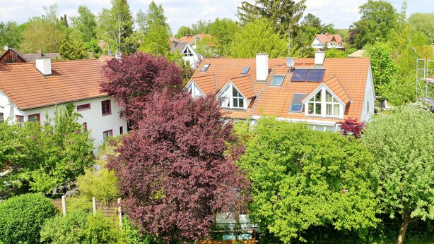 Ein Wohngebiet mit Häusern, die roten Ziegeldächer und Solaranlagen haben, sowie einer Mischung aus Laubbäumen.