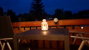 Ein Tisch mit einer Laterne, umgeben von Stühlen, draußen mit Holzlattenzaun und Bäumen im Hintergrund.