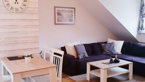 Ein gemütliches Wohnzimmer mit einer Holzwand, einem dunklen Sofa, einem weißen Tisch und einer Uhr mit römischen Ziffern.