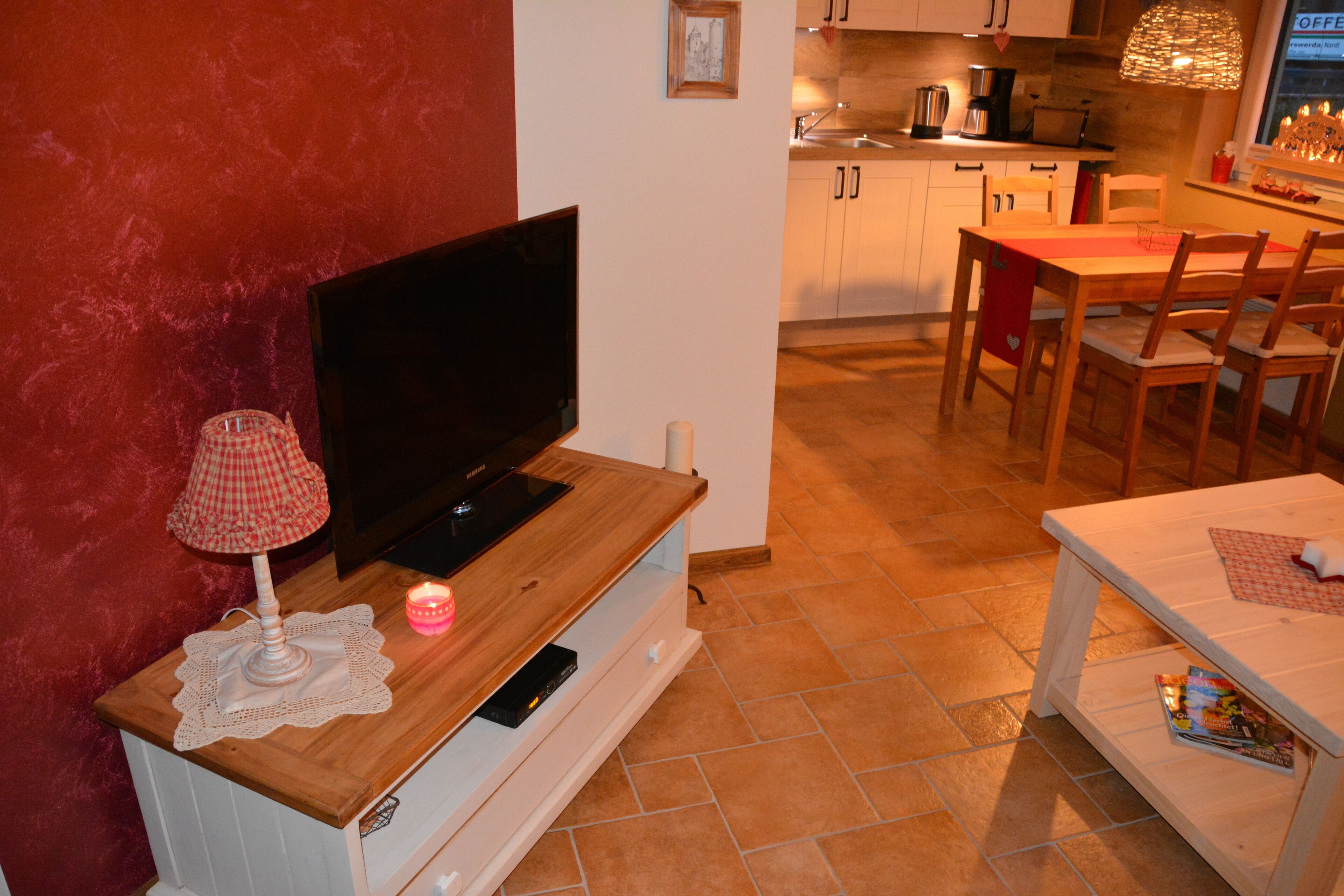 A living room with a television on a wooden stand, a small table with a lamp, and a dining area with chairs and a table.