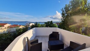 Une terrasse sur le toit, dotée d’un espace salon comprenant deux chaises et une petite table, offrant une vue sur un paysage urbain côtier avec ses bâtiments et un ciel dégagé.