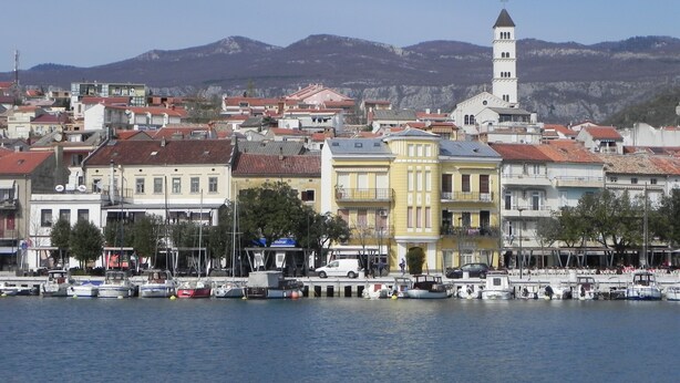 Une ville côtière dotée d’une tour imposante, de bâtiments aux couleurs vives et d’un port de plaisance où sont amarrés des bateaux.