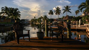 A wooden deck with two chairs and a potted plant, overlooking a marina with boats and palm trees. - Dream villa in top location on the wide canal & large pool area with jacuzzi (Cape Coral)